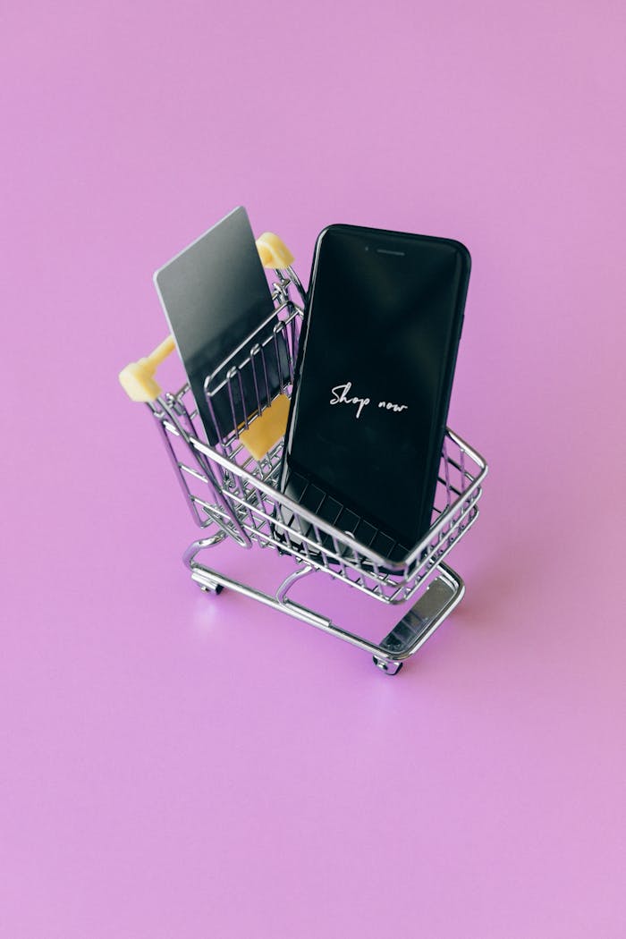 Modern shopping concept featuring smartphone and credit cards in a miniature cart on pink background.
